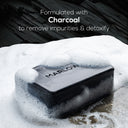 Marlowe charcoal exfoliating soap bar with natural ingredients, showing the dark soap with textured surface and herbal inclusions.